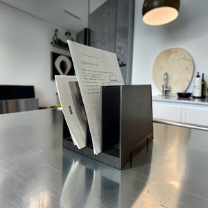 May include: A matte black metal desktop file organiser with three vertical compartments. The organiser holds several white papers with handwritten notes and a small printed document with the text "MIMICA" visible on the lower left corner.