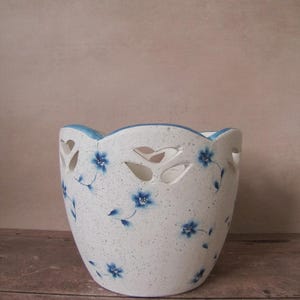 May include: A white ceramic planter with a speckled finish and a scalloped rim. The planter features cut-out leaf shapes and is decorated with blue floral patterns. The rim is painted blue. The planter sits on a wooden surface.