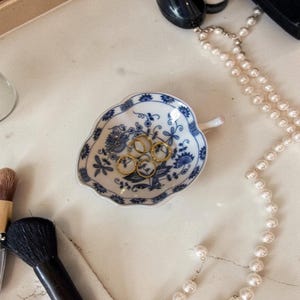 Puede incluir: Un pequeño cuenco en forma de hoja con un diseño floral azul y blanco contiene tres anillos de oro. Un collar de perlas y brochas de maquillaje también son visibles sobre una superficie blanca. El cuenco es decorativo.