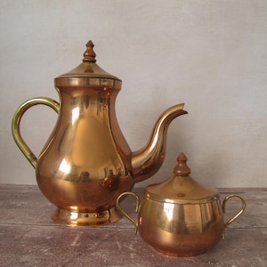 Peut inclure: Un service à thé en cuivre comprenant une théière et un sucrier. La théière a un bec verseur incurvé et un bouton en bois sur le couvercle. Le sucrier a deux poignées et un bouton en bois assorti. L'ensemble est sur une surface en bois.