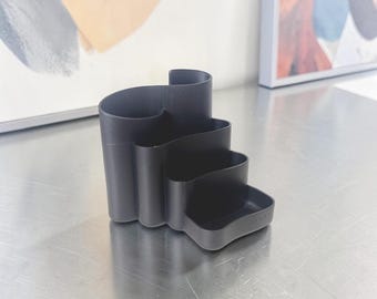 May include: A black desk organizer with a unique, tiered design. The organizer has multiple compartments of varying heights and depths, perfect for storing pens, pencils, and other office supplies. The organizer is made of a matte black material.