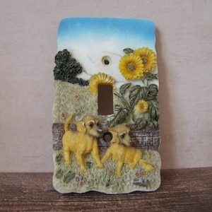 Puede incluir: Cubierta decorativa para interruptor de luz que representa una escena campestre con dos perros amarillos en un campo de hierba, girasoles y un cielo azul. La cubierta tiene forma rectangular con un hueco para el interruptor.