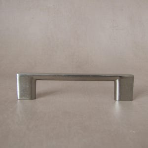 Op de afbeelding: Een zilverkleurige kastgreep met een rechthoekige stang en vierkante uiteinden. De metalen hardware is ontworpen voor laden of kastdeuren. De greep wordt getoond tegen een neutrale achtergrond. Ongeveer 10 cm lang.