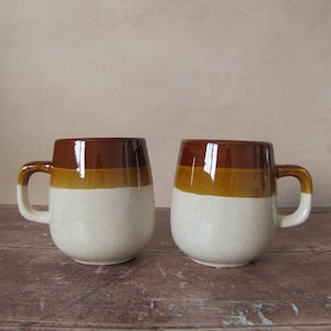 Puede incluir: Dos tazas de cerámica con forma redondeada. Cada taza presenta una parte superior marrón, una sección media beige y una base blanca moteada. Las asas son blancas con un acento marrón. Las tazas están sobre una superficie de madera.