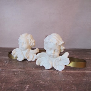 Könnte beinhalten: Zwei cremefarbene Cherubinskulpturen mit detaillierten Flügeln und lockigem Haar ruhen auf einem goldfarbenen Band. Die Cherubim sind auf einer dunkelbraunen Holzoberfläche platziert, was einen Kontrast erzeugt.