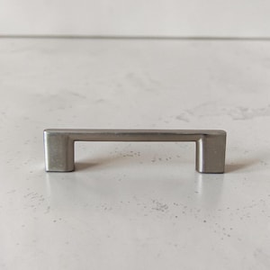 Op de afbeelding: Een greep van geborsteld nikkel met een rechthoekige stang en twee vierkante stijlen. De hardware is ontworpen voor eenvoudige installatie en voegt een moderne touch toe aan laden en kasten. De greep is ongeveer 10 cm lang.