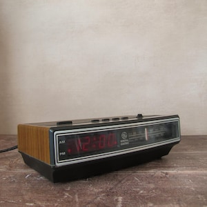 Op de afbeelding: Vintage General Electric digitale wekker met een houtnerf afwerking aan de zijkanten. De klok geeft de tijd weer in rode cijfers en heeft een zwart en zilveren voorkant. Geplaatst op een houten oppervlak.