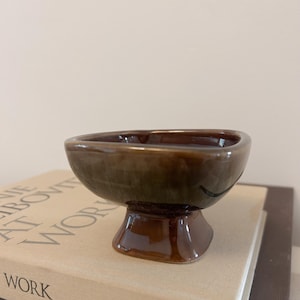 May include: A small, brown ceramic bowl with a pedestal base. The bowl has a square shape with rounded corners and a dark brown interior. The exterior has a glossy, mottled finish with shades of brown and green.