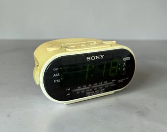 Könnte beinhalten: Ein blassgelber Sony Dream Machine Radiowecker mit schwarzer Frontblende. Das digitale Display zeigt die Uhrzeit in Grün an, inklusive AM-, PM- und DST-Anzeigen. Die Vorderseite enthält FM- und AM-Radiofrequenzskalen sowie Alarmeinstellungen.