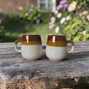 Puede incluir: Dos tazas de cerámica con forma redondeada y asas pequeñas. Las tazas están divididas en tres secciones horizontales: una base moteada blanquecina, una banda de color amarillo dorado y una parte superior marrón oscuro. Se encuentran sobre una superficie de madera envejecida.
