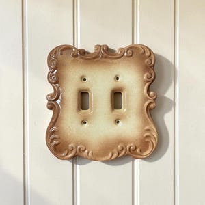 May include: A double light switch wall plate featuring a glossy, cream-coloured centre that fades into a darker tan, scalloped, and embossed border with intricate scrollwork details. The rectangular plate includes two vertical toggle switch openings.