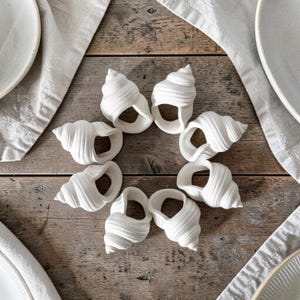 May include: Eight white seashell-shaped napkin rings are arranged in a circle on a wooden surface. The rings are made of a smooth, ceramic-like material and are designed to hold cloth napkins. White plates and napkins are in the background.