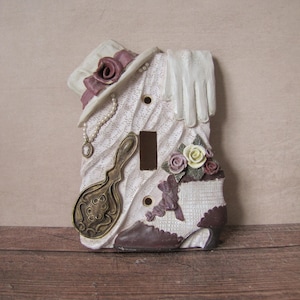 Puede incluir: Una cubierta decorativa para interruptor de luz con un diseño vintage. La cubierta presenta un sombrero, un guante, un espejo y un zapato de tacón alto, en tonos crema y marrón. Incluye un collar de perlas y detalles florales, creando una estética victoriana.