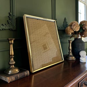 May include: A rectangular picture frame with a thin, polished gold-tone metal border and a clear glass front. The frame is empty, revealing a tan-coloured backing material inside. The design features a simple, clean edge with a subtle textured pattern.