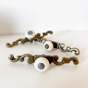 Może przedstawiać: Trzy dekoracyjne uchwyty do szafek o estetyce vintage. Każdy uchwyt posiada nakrapiane białe ceramiczne pokrętło z szarym środkiem, osadzone na podstawie z metalu w odcieniu brązu z ozdobnymi, zakrzywionymi detalami. Uchwyty są ułożone na białej powierzchni.