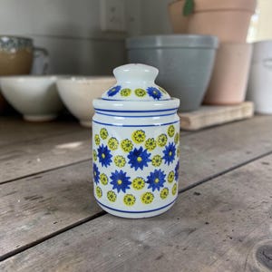 Peut inclure: Petit pot en céramique blanche avec couvercle, orné d'un motif répété de fleurs stylisées bleues et jaunes. Le pot est cylindrique avec un dessus arrondi et repose sur une surface en bois. Le couvercle reprend le motif floral.