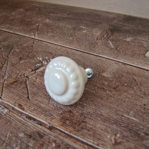 Może przedstawiać: Białe ceramiczne pokrętło do szafki z dekoracyjnym, żebrowanym wzorem. Pokrętło ma mały, przezroczysty szklany akcent. Pokrętło jest umieszczone na rustykalnej, brązowej drewnianej powierzchni.