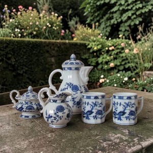 May include: A white porcelain tea set with blue floral patterns. The set includes a teapot, creamer, sugar bowl, and two small mugs. The pieces are arranged on a weathered wooden surface, with a garden in the background.