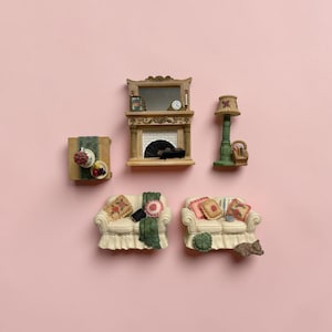 Op de afbeelding: Een miniatuur woonkamerset bestaande uit twee crèmekleurige banken met bloemenkussens en groene plaids, een open haard met spiegel, klok en fotolijst, een vloerlamp en een vierkante tafel met een kleine schaal met fruit en bloemen.