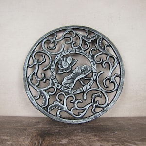 May include: Circular cast iron trivet with an ornate scrolling design. The centre features a floral motif within a circular frame. The trivet has a distressed silver and black finish, adding a vintage aesthetic. This decorative item is designed to protect surfaces from heat.