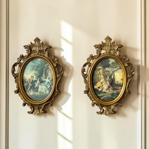 May include: Two oval wall frames with ornate gold-coloured detailing and scrollwork. Each frame contains a printed pastoral scene featuring figures in outdoor settings with trees and greenery. The frames have a decorative top and bottom crest.