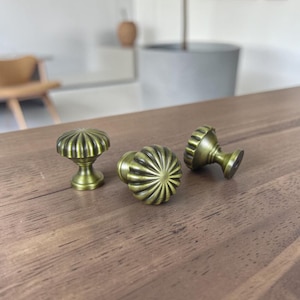 Könnte beinhalten: Drei antike Messing-Schrankknöpfe mit geripptem, sonnenblumenartigem Design liegen auf einer Holzoberfläche. Die Knöpfe haben eine Vintage-Ästhetik mit einem warmen, gealterten Messing-Finish. Sie sind für Möbelrestaurierung oder Heimdekoration.