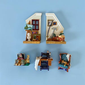 May include: A collection of five miniature resin figurines including two wall-shaped room sections with art supplies and a bicycle, a toy box with a clown and teddy bear, a vintage sewing machine with white fabric, and an armchair with a sleeping cat.