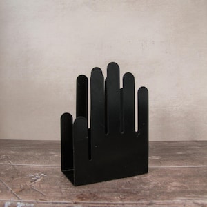Puede incluir: Un servilletero de metal negro con forma de mano abierta. El soporte está hecho de una sola pieza de metal con dedos recortados. Se encuentra sobre una superficie de madera y tiene un diseño minimalista.