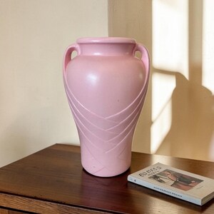 May include: A matte light pink ceramic vase featuring two integrated side handles and a decorative embossed crisscross pattern on the lower body. The vessel has a wide, rounded rim and a tapered base, presenting a smooth, uniform finish.
