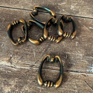 May include: A collection of six antique bronze-colored drawer pulls. Each pull has a curved, oval shape with a decorative, ridged handle. The pulls are arranged on a weathered, wooden surface, showcasing their vintage aesthetic.