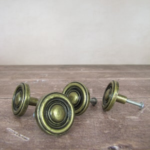 May include: Four antique brass cabinet knobs with a circular design. The knobs have a dark, aged finish and are arranged on a wooden surface. The knobs are likely for furniture restoration or decorative purposes.