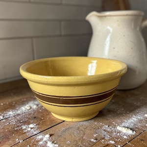 May include: A yellow ceramic mixing bowl with two brown stripes. The bowl sits on a wooden surface with flour scattered around it. A cream-coloured jug and a wooden chopping board are in the background.