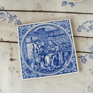 Può includere: Piastrella quadrata in ceramica blu e bianca con illustrazione storica. L'immagine raffigura una scena con figure in una stanza con scaffali, incorniciata da un bordo floreale. Il testo include "De Advokaat" e le date 1649-1713.