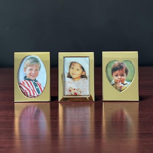 May include: Three gold-toned metal picture frames standing in a row. The left frame has an oval opening, the center frame has a rectangular opening, and the right frame has a heart-shaped opening. Each frame contains a photograph of a young child.