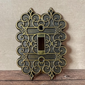 Puede incluir: Cubierta de interruptor de luz ornamental de color bronce con un diseño decorativo de volutas. La cubierta tiene una abertura rectangular para el interruptor y está montada sobre una superficie de madera. El diseño presenta remolinos y florituras simétricas.