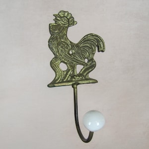 Könnte beinhalten: Ein dekorativer Wandhaken mit einem detaillierten, messingfarbenen Hahn-Design. Der Haken hat einen gebogenen, dunkelmetallenen Stiel und einen weißen, runden Knopf am Ende. Dieser Haken im Vintage-Stil eignet sich für Küche oder Eingangsbereich.