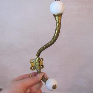 Peut inclure: Un crochet mural vintage avec une finition en laiton et des boutons en céramique blanche. Le crochet présente un design torsadé décoratif et un motif floral sur la plaque de fixation. Longueur totale d'environ 18 cm.