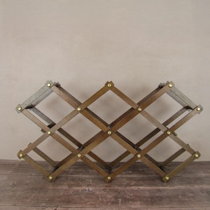 Könnte beinhalten: Ein Weinregal aus Holz mit rautenförmigem Design. Das Regal besteht aus dunkelbraunem Holz und hat messingfarbene Nieten an jeder Verbindung. Es ist für mehrere Weinflaschen konzipiert und wird auf einer Holzoberfläche präsentiert.