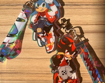 May include: Two colorful keychains featuring cartoon characters Sonic and Shadow. Both characters are depicted in a street style aesthetic with red, blue, and black clothing. The keychains are attached to iridescent straps with the words "Sonic" and "Shadow" printed on them.