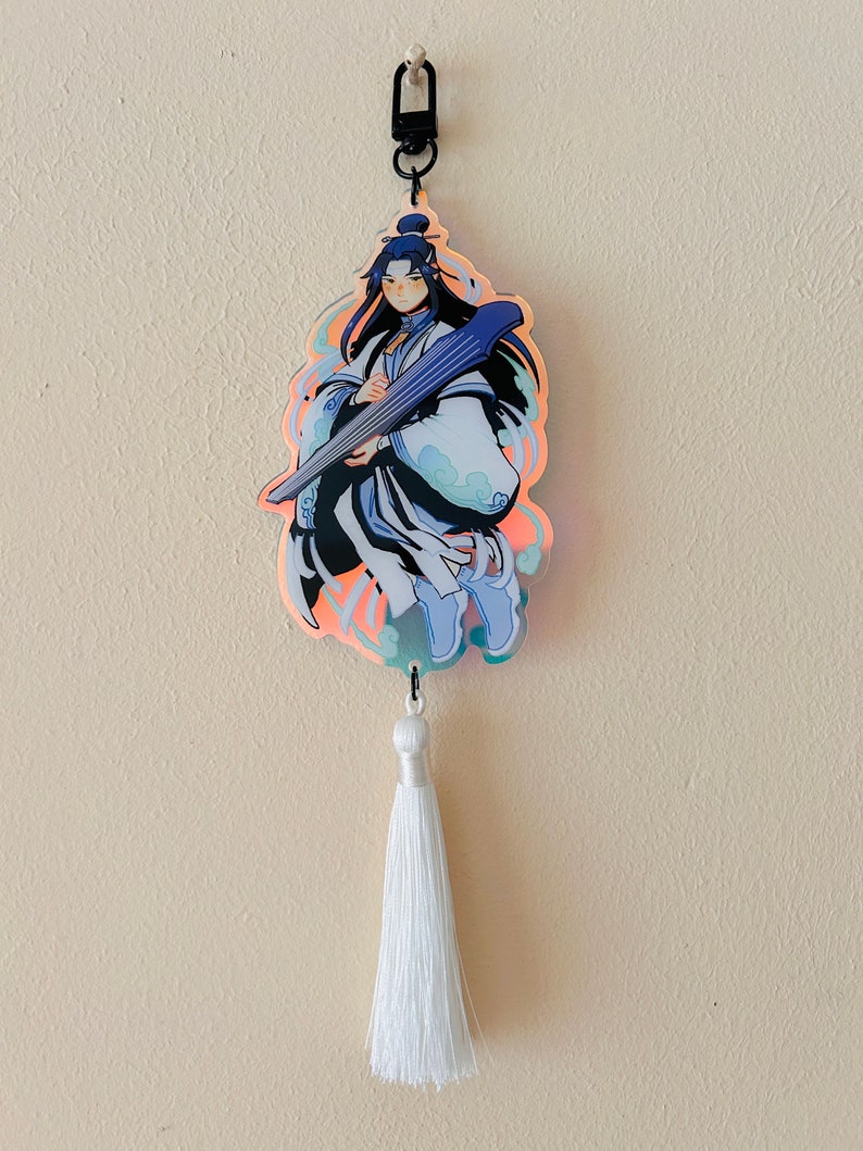 May include: A holographic charm featuring a character with long black hair holding a blue and white instrument. The charm is attached to a white tassel.