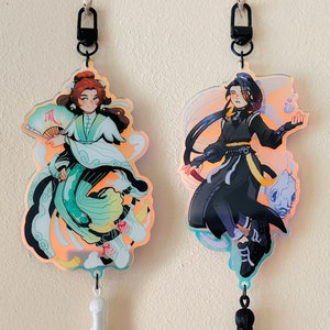 May include: Two colorful anime-style character charms with black keychains. The charm on the left features a character with long brown hair, wearing a green and white outfit, holding a fan with the word "风" on it. The charm on the right features a character with long black hair, wearing a black and white outfit, holding a white object. Both charms have a holographic effect.