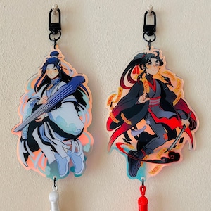 May include: Two iridescent keychains featuring anime characters. The keychain on the left features a character with blue hair playing a string instrument. The keychain on the right features a character with black hair holding a sword. Both keychains have red and gold accents.