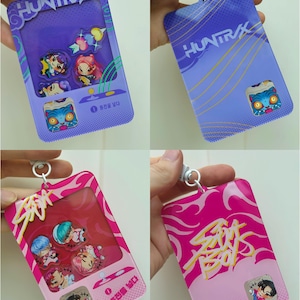 Könnte beinhalten: Vier Schlüsselanhänger im Bild. Zwei sind lila mit dem Text "HUNTRAX", und zwei sind rosa mit dem Text "SOUL BOYS". Jeder Schlüsselanhänger hat ein klares Fenster mit Cartoon-Charakter-Designs. Die Schlüsselanhänger haben einen silbernen Ring zur Befestigung.