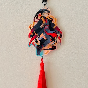 May include: A red tassel keychain with a clear acrylic charm featuring an anime-style character with black hair, a black and red outfit, and a sword. The character is surrounded by gold and red swirls.