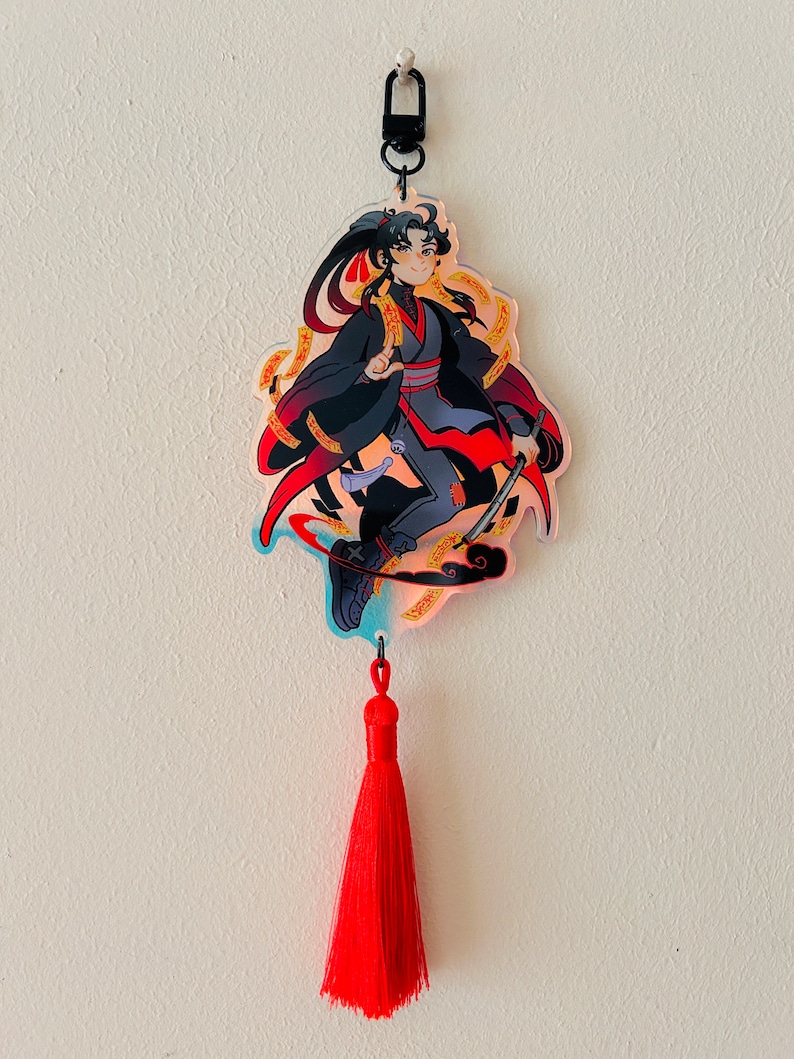 May include: A red tassel keychain with a clear acrylic charm featuring an anime-style character with black hair, a black and red outfit, and a sword. The character is surrounded by gold and red swirls.