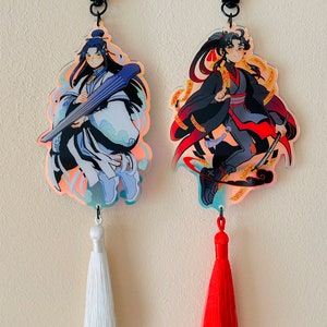 May include: Two anime-style character keychains with tassels. The keychain on the left features a character with long blue hair playing a string instrument. The keychain on the right features a character with black hair holding a sword. Both keychains have a holographic effect.