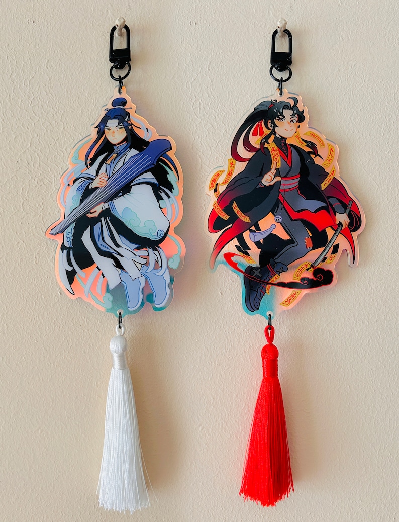 May include: Two anime-style character keychains with tassels. The keychain on the left features a character with long blue hair playing a string instrument. The keychain on the right features a character with black hair holding a sword. Both keychains have a holographic effect.