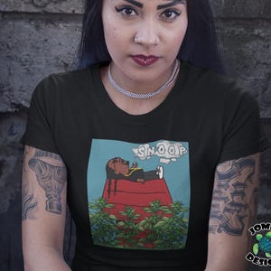 May include: Black t-shirt featuring a cartoon illustration of Snoop Dogg relaxing on a red doghouse, with the word "SNOOP" above. The design includes green plants and a blue sky background. The shirt is a crew neck.