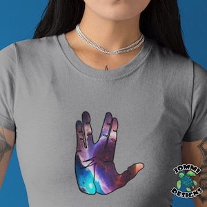 Peut inclure: Un t-shirt gris avec un geste de salutation vulcanien à thème galactique. La main est de couleur bleu foncé, violet et rose avec une étoile blanche au centre. Le texte "JOMMI DESIGNS" est dans le coin inférieur droit de l'image.