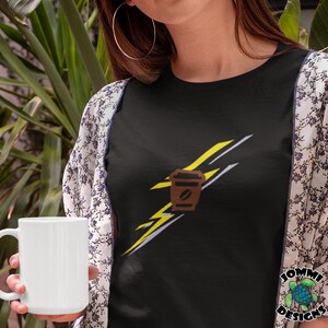 以下が含まれることがあります： 黄色の稲妻のグラフィックと茶色のコーヒーカップのデザインが施された黒いTシャツ。画像の右下隅に「JOMMI DESIGNS」というテキストが表示されています。